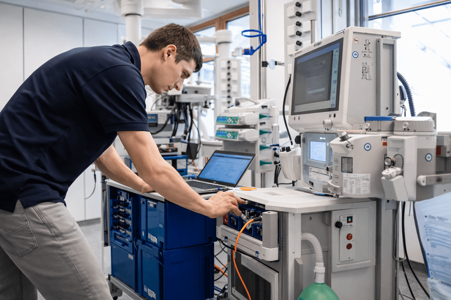 Technician in a blue shirt inspects medical equipment using a laptop in a clinical laboratory.