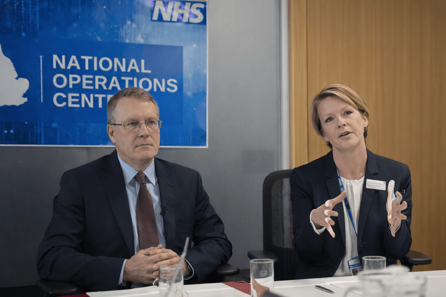 Woman gesturing while speaking beside a man at the NHS National Operations Centre.