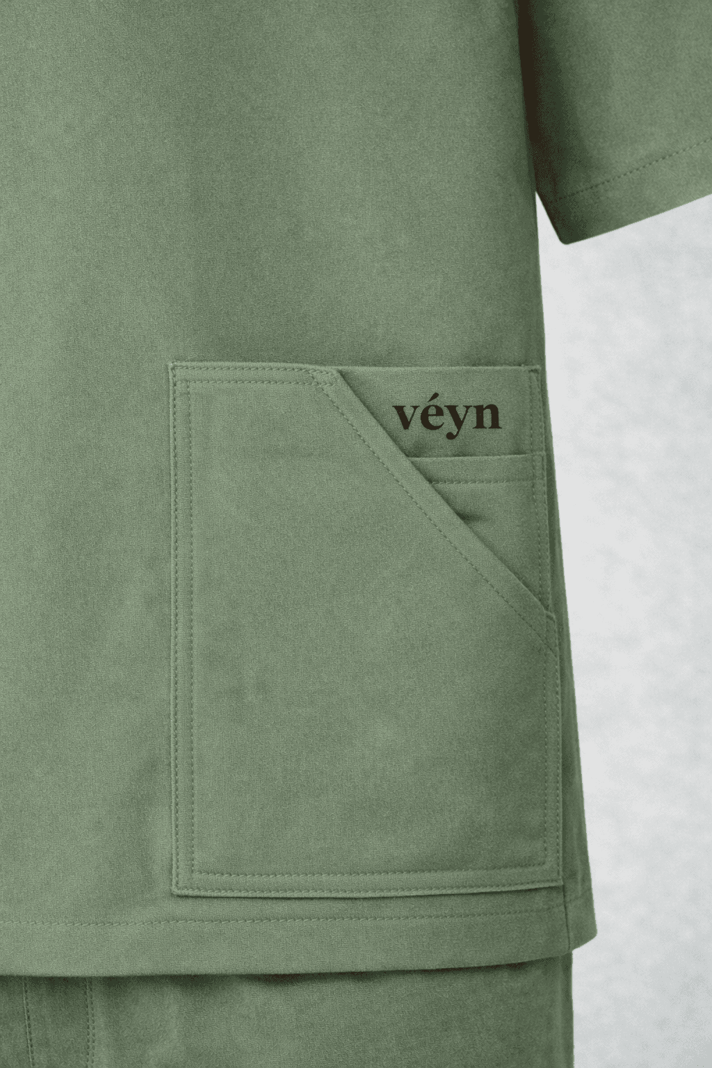 Close-up of sage green scrubs with a large pocket and red embroidered "véyn" logo.